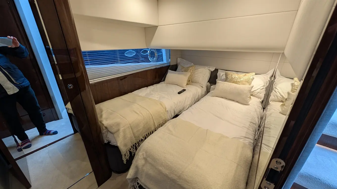 Bright, modern cabin featuring two twin beds with white linens, gold cushions, and a remote on one bed. Natural light enters through blinds.